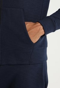 Felpa zip con cappuccio di colore blu navy realizzata in tessuto morbido, con una tasca sul lato destro, bordi a costine e una texture liscia.