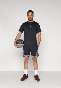 Μαύρο αθλητικό μπλουζάκι με λογότυπο, μαύρο σορτς με λευκή επένδυση και μαύρα-κόκκινα αθλητικά παπούτσια. Κρατώντας μια μπάλα μπάσκετ με λογότυπο.