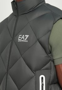 Vesta puffer neagră cu model diamantat, guler înalt și buzunar lateralt cu fermoar. Prezintă logo-ul EA7 Emporio Armani în alb.