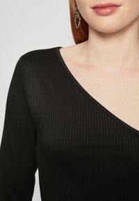 Černý žebrovaný top s hlubokým výstřihem do V a dlouhými rukávy. Obsahuje jemnou texturu a hladký povrch. Doplněno náušnicí ve tvaru srdce.
