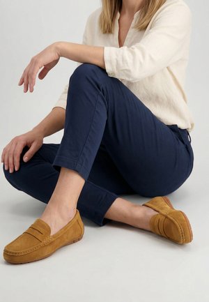 Vrouw zittend met gekruiste benen, draagt marineblauwe broek, crèmewitte lange-mouwen blouse en beige suède loafers op een witte achtergrond.