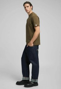 T-shirt verde oliva a maniche corte, abbinata a jeans di jeans scuro e stivali neri. La t-shirt presenta un piccolo logo sulla manica.