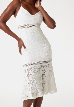 Vue en longueur d’une femme portant une robe midi blanche en dentelle fleurie avec des bretelles spaghetti, des panneaux transparents à la taille et à l’ourlet, posant avec la main sur la hanche.
