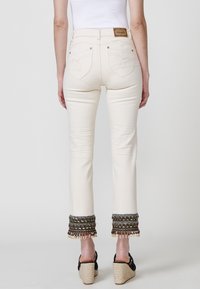 Pantalones de mezclilla blanca con pierna acampanada, que presentan dobladillos adornados con intrincados bordados y borlas, y bolsillos traseros con costuras decorativas.