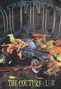 Černé tričko s barevným grafickým designem, včetně plamenů a automobilu, s výrazným modrým a žlutým textem "OUTUT" a "THE COUTURE CLUB."