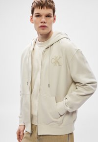 Bézs zipzáras kapucnis pulóver elülső zsebbel, húzózsinórokkal és hímzett logóval. Krémszínű bordázott pulóver fölött viselve, világos bézs nadrággal párosítva.