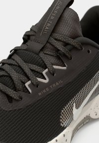 Gros plan d'une chaussure de trail Nike noire avec tissu texturé, lacets à motifs, logo swoosh blanc et détails de semelle mouchetée.