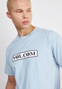 Camiseta de algodón de color azul claro que presenta un logo rectangular blanco con borde negro, con el texto "VOLCOM" y un emblema centrado sobre él.