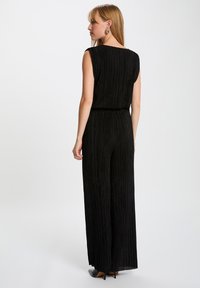 Zwarte jumpsuit met verticale ribbelstructuur, mouwloos ontwerp, wijde pijpen en een cinched taille. Gestyld met hakken en minimale sieraden.