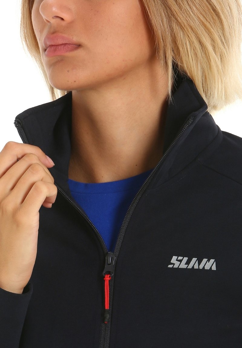Veste zippée marine avec col montant, ornée d'un logo argenté bien en vue et d'une fermeture éclair rouge, portée par-dessus une chemise bleue. Texture de tissu lisse.