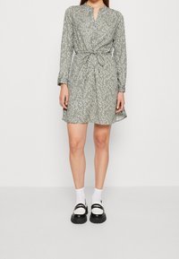 Vestido floral verde con mangas largas, un lazo en la cintura y una fila de botones. Combinado con calcetines blancos y zapatos gruesos en blanco y negro.