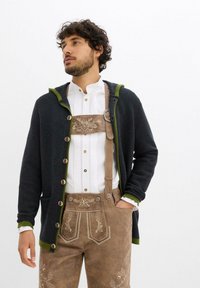 Cardigan lavorato a maglia di colore blu scuro con dettagli verdi, dotato di grandi bottoni e cappuccio, indossato sopra una camicia bianca e pantaloni lederhosen marroni ricamati.