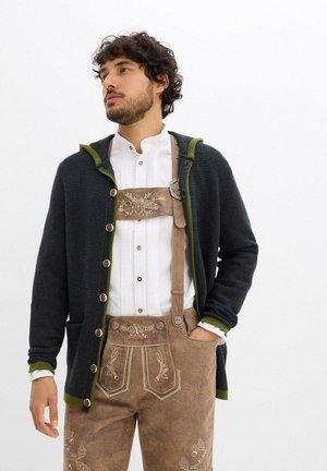 Cardigan en tricot bleu foncé avec des accents verts, présentant de grands boutons et une capuche, porté sur une chemise blanche et un lederhosen brodé marron.