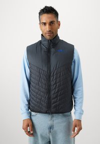 Padded vest w ciemnoszarym kolorze z pikowanym wzorem, wysokim kołnierzem i niebieskim logo "INTER". W połączeniu z jasnoniebieską koszulą z długim rękawem.