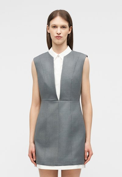 Γυναίκα με ίσια καστανά μαλλιά, φοράει αμάνικο γκρι φόρεμα με βαθύ V ντεκολτέ πάνω από λευκό πουκάμισο με γιακά, στέκεται μπροστά σε απλό φόντο.