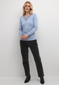 Žena nosi svijetloplavu bluzu dugih rukava s čipkastim V-izrezom, crne traperice visokog struka i crne čizme s petom iznad gležnja, stoji uz bijeli zid.