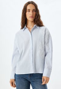 Camisa de botones de rayas azul claro con un corte relajado. Presenta un detalle floral bordado en un lado y puños a rayas en contraste.