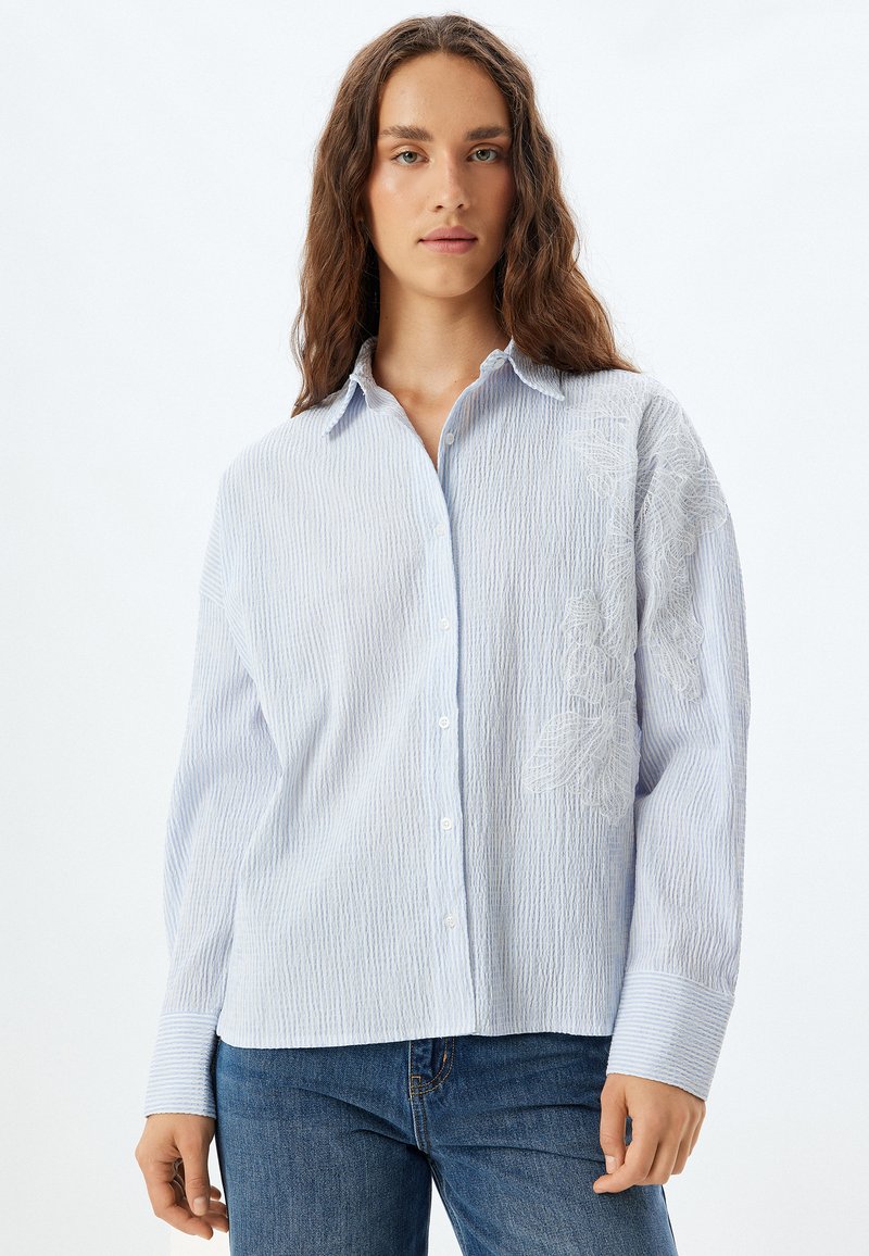 Camisa de botones de rayas azul claro con un corte relajado. Presenta un detalle floral bordado en un lado y puños a rayas en contraste.