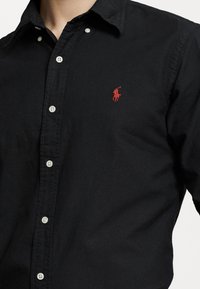 Czarna koszula z długim rękawem, wykonana z bawełny, z kołnierzykiem i białymi guzikami. Widoczny czerwony logo polo na lewej piersi. Gładka tekstura.