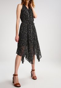 Bezramionna czarna sukienka z kwiatowym wzorem, z wcięciem w talii i asymetrycznym dołem. Połączona z brązowymi sandałami na słupku.