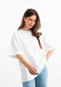 T-shirt bianco oversize in morbido tessuto, con testo ricamato blu "MIRONS", maniche corte e vestibilità rilassata, abbinato a jeans blu chiaro.