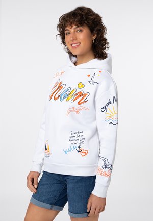 Junge Frau mit lockigem Haar, die einen weißen Hoodie mit buntem „Moin“-Schriftzug, nautischen Motiven und deutscher Schrift trägt, kombiniert mit Jeansshorts.