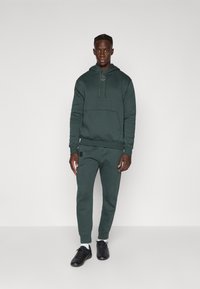 Felpa color verde scuro con pantaloni abbinati, realizzati in tessuto morbido. La felpa presenta una tasca a marsupio e un cordoncino, con dettagli del logo. Sneakers nere.