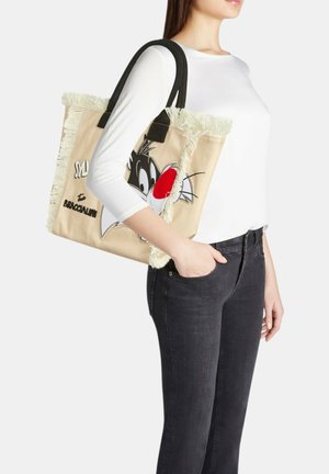 Donna che porta una borsa a tracolla beige in tela con manici neri, bordi sfrangiati e una faccia di gatto in stile cartone animato con un naso rosso di lato.