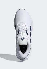 Chaussure de sport blanche avec des bandes bleu marine, dessus en mesh et lacets, vue de dessus sur un fond clair.
