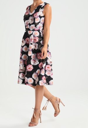 Mujer con vestido midi negro sin mangas con estampado floral en rosa y blanco, combinado con sandalias de tacón alto color nude con lazos en el tobillo.