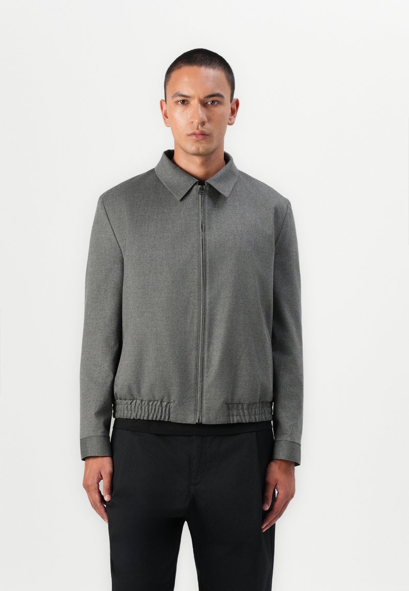Jachetă gri cu fermoar, cu guler, tiv elastic și mâneci lungi. Textură netedă, design ajustat și stil minimalist.