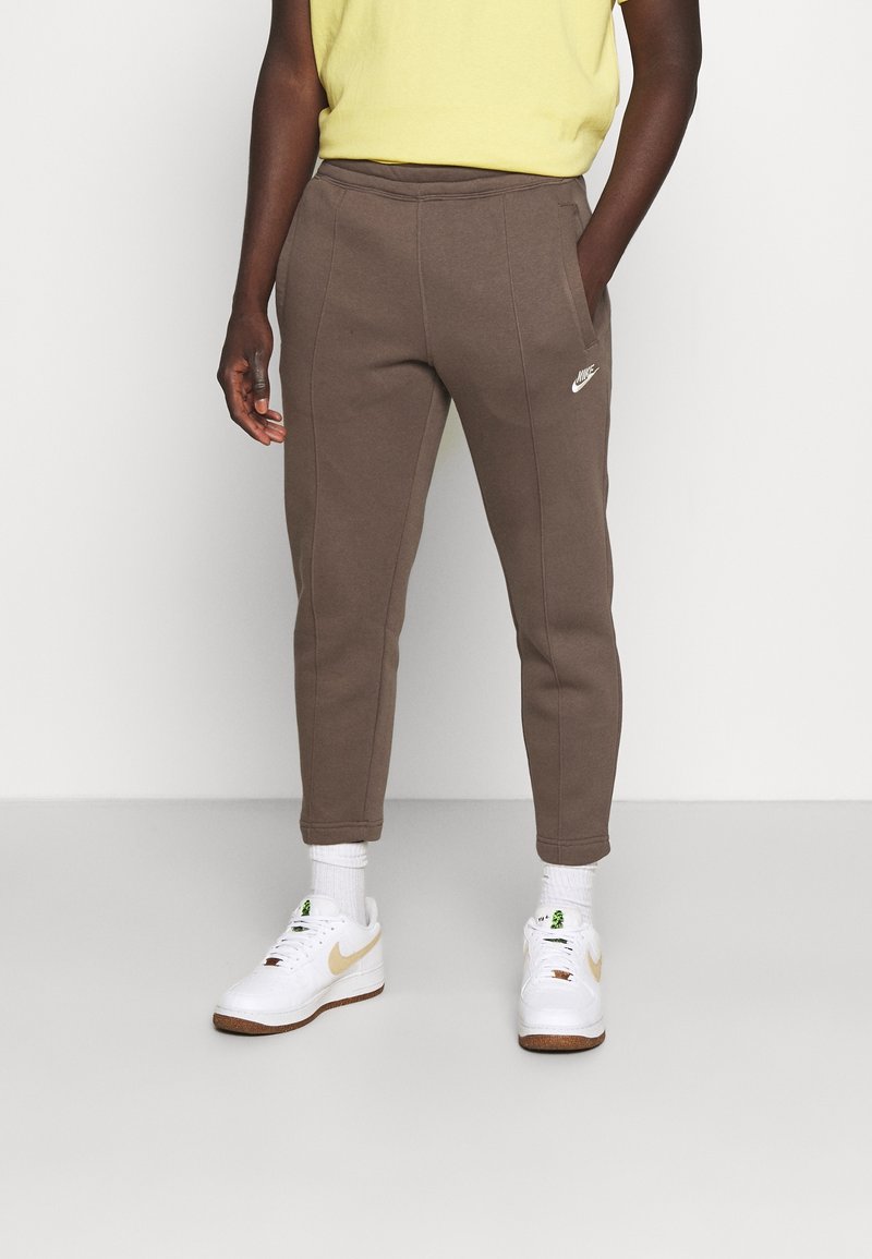 Hnědé tepláky se zúženým střihem, elastickým pasem, bočními kapsami a bílým logem Nike. V kombinaci s bílými teniskami a kotníkovými ponožkami.