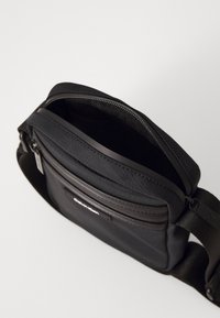 Borsa a tracolla nera con tessuto testurizzato, chiusura con zip e tracolla regolabile. Presenta una tasca frontale con etichetta del logo del brand.