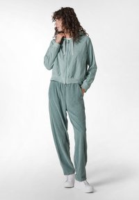 Chaqueta verde clara con cremallera y capucha, textura acanalada y cordones, combinada con pantalones ajustados a juego, ambos con bolsillos laterales.