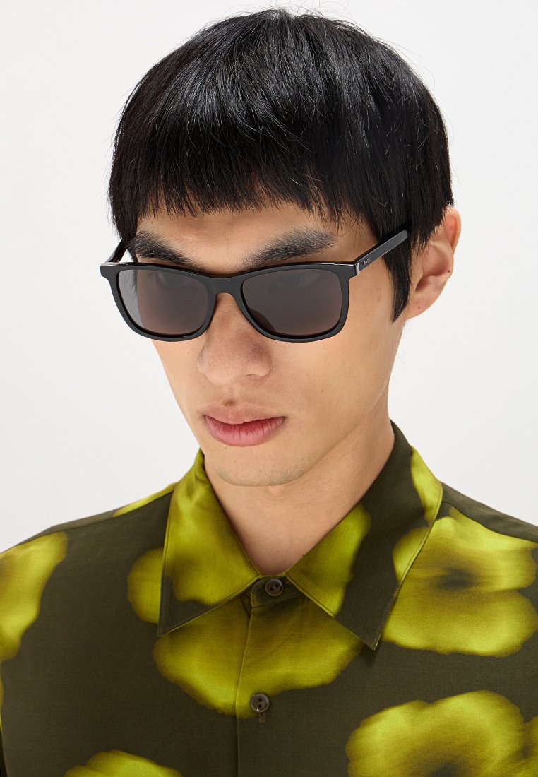 Schwarze rechteckige Sonnenbrille mit dunklen Gläsern. Das Modell trägt ein grünes Shirt mit großen gelben Blumenmustern. Weicher Stofftextur.