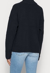 Pull côtelé bleu marine avec un col montant et des épaules tombantes, doté d'une coupe décontractée et d'un tricot texturé. Porté avec un jean bleu clair.