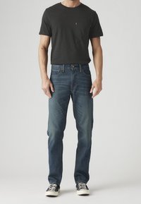Homme portant un t-shirt noir à manches courtes, un jean bleu et des baskets noires, debout devant un fond clair uni.