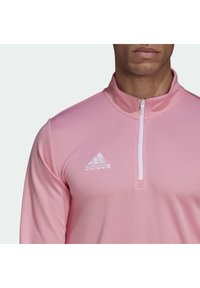 Rosa idrottströja med dragkedja, vit dragkedja, hög krage och broderad Adidas-logotyp på vänster bröst. Mjuk, slät tygkvalitet.