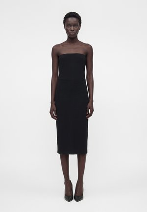 Femeie în picioare, purtând o rochie midi neagră, mulată, fără bretele și pantofi negri cu vârf ascuțit, pe un fundal alb simplu.