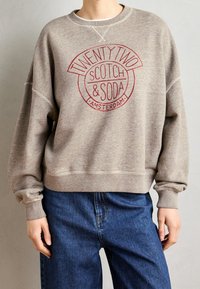 Sudadera beige con logo circular en rojo que dice "TWENTY TWO SCOTCH & SODA AMSTERDAM." Presenta un escote amplio y un ajuste holgado.
