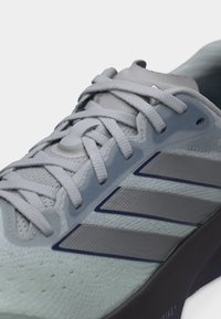 Ανοιχτό γκρι παπούτσι τρεξίματος με τρεις σκούρες γκρι ρίγες, λευκά κορδόνια και μαύρη μαξιλαρωμένη σόλα.