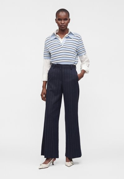 Femme debout portant un pull à manches courtes rayé bleu clair et bleu marine sur une chemise blanche à col, un pantalon large à rayures bleu marine et des talons blancs.