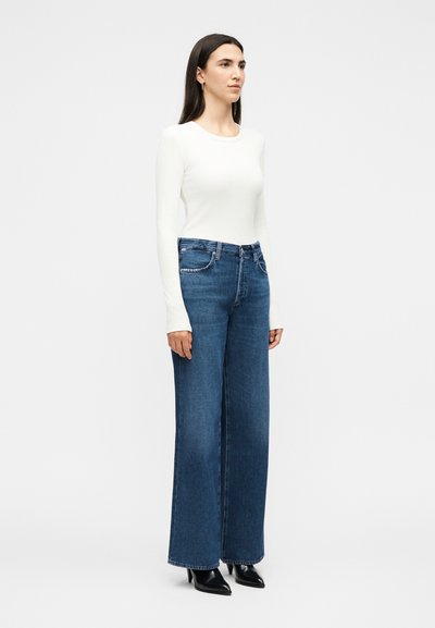 Γυναίκα με μακριά σκούρα μαλλιά, ντυμένη με λευκό μπλουζάκι μακρυμάνικο, φαρδιά μπλε τζιν παντελόνια και μαύρα μυτερά παπούτσια με τακούνι, στέκεται μπροστά σε λευκό φόντο.