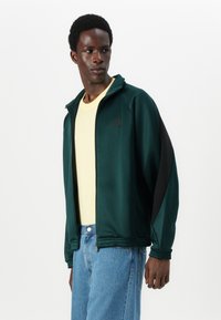 Πράσινο τζάκετ με φερμουάρ και μαύρες λεπτομέρειες, φτιαγμένο από λείο ύφασμα. Φοριέται πάνω από ένα ανοιχτό κίτρινο πουκάμισο. Το μοντέλο φοράει μπλε τζιν.