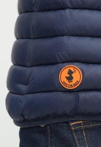 Tamnoplavi perjani kaput s narančastim okruglim oznakom na kojoj je crna patka i natpis "SPASITE PATKU" iznad plavih traperica.