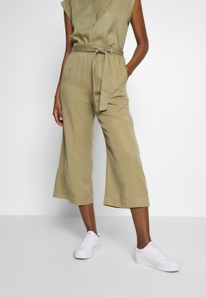 Person, die olivgrüne, weit geschnittene, gekürzte Hose mit gebundenem Taillengürtel und weißen Turnschuhen trägt, steht vor weißem Hintergrund.