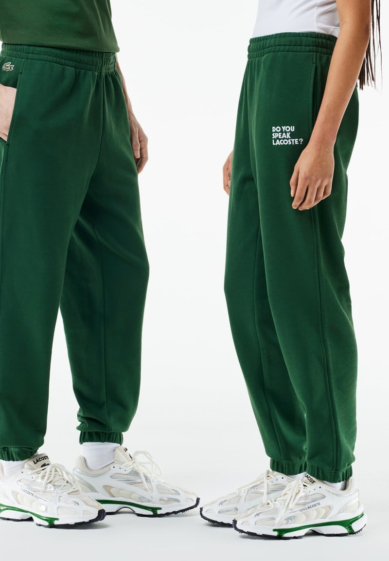 Zielone spodnie dresowe z elastycznym pasem i mankietami. Na udzie znajduje się biały napis "CZY MÓWISZ LACOSTE?". Noszone z białymi trampkami.