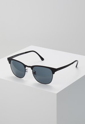 Occhiali da sole rettangolari con montatura nera e lenti scure appoggiati su un cubo bianco su uno sfondo grigio.