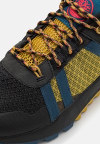 Gros plan sur une chaussure de randonnée avec un mesh jaune, un tissu noir, des accents bleus et des lacets à motifs jaune-noir-rouge.