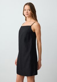 Robe en lin noire avec un encolure carrée et des fines bretelles. Le design est simple, sans ornements, et arrive au-dessus du genou.
